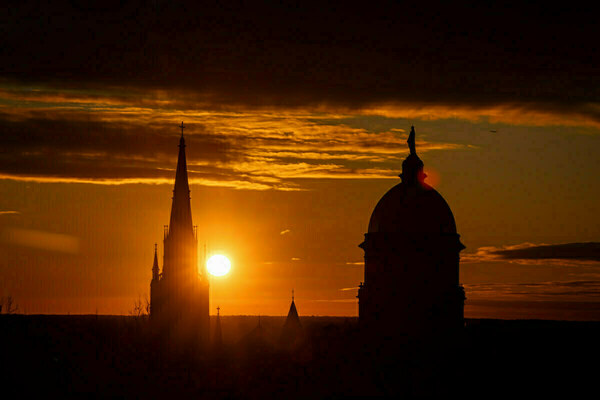 The Basilica of the Sacred Heart steeple and the Main Building stand silhouetted against a brilliant orange and yellow sunset. The bright sun glows behind the steeple, with colorful clouds above.