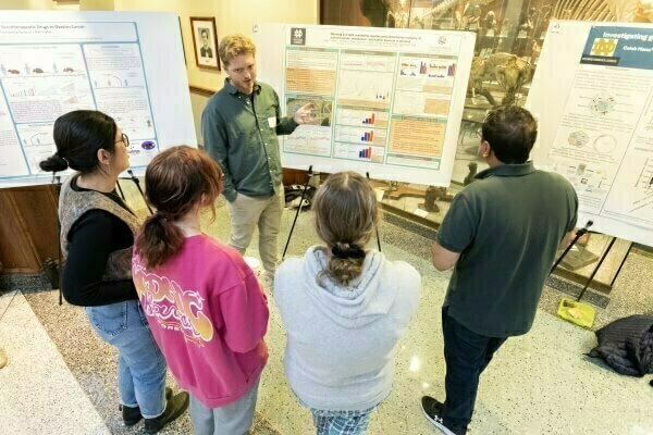 A male presenter in a green shirt gestures at a scientific poster with a Notre Dame logo to four students. Visible from behind, they include two women in hoodies (pink, gray), a woman in a vest, and a man in a polo, attentively observing in a building with museum exhibits.