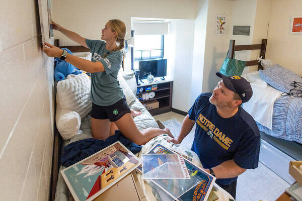 Blonde woman in teal shirt hangs art on dorm wall. Man in Notre Dame hat & shirt watches, helping move in.